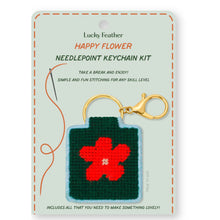 Load image into Gallery viewer, This image shows a needlepoint keychain kit packaged on a light sage-green card featuring a decorative brown stitched border. The card displays a finished sample consisting of a dark green woven square with a bright red flower charm attached to a gold key ring and clasp, next to a graphic illustration of a sewing needle.
