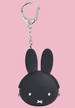 Load image into Gallery viewer, This is a small, round silicone coin purse keychain shaped like the head of the cartoon rabbit Miffy, set against a solid pink background. The pouch features a smooth black face with minimalist white details: two small dot eyes and a tiny "X" shaped mouth. Two long, rounded ears extend straight up from the top. A silver ball chain links the purse to a carabiner-style key clip. It uses a metal clasp hidden within the silicone frame, showing only the silver hinges on the sides.
