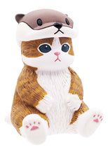 Load image into Gallery viewer, A small brown-tabby cat figurine sitting upright against a plain white background, wearing a sea otter hood on its head. The hood sits like a cap, featuring a simple otter face with black eyes and a dark nose resting just above the cat's wide, wide-eyed gaze and pink-padded feet.
