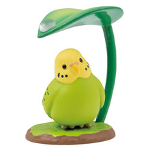 Load image into Gallery viewer, A round green parakeet figurine with a bright yellow head, sitting on a small patch of grass against a plain white background. The bird tilts its head slightly under a large green leaf that curves over it like an umbrella, bearing a single clear raindrop on top.

