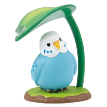 Load image into Gallery viewer, A round, vibrant blue parakeet figurine with a white head and yellow beak, sitting on a small patch of grass against a plain white background. The bird tilts its head curiously under a curved green leaf that serves as an umbrella, decorated with a single clear raindrop on top. 
