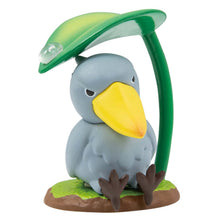 Load image into Gallery viewer, A blue-grey shoebill bird figurine sitting on a grassy base against a plain white background, its dark feet splayed forward. The bird has a large, prominent yellow beak and an intense gaze beneath a tall green leaf stem that arches over its head like an umbrella, bearing a single clear raindrop.
