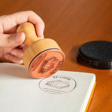 Load image into Gallery viewer, This image shows a person holding the wooden handle of the book stamp, hovering it over a crisp, black circular imprint freshly stamped onto the blank white page of an open notebook. The imprint reads "EX-LIBRIS" over a graphic of an open book with space below to write a name, while the uncovered black ink pad sits nearby on the smooth wooden table.
