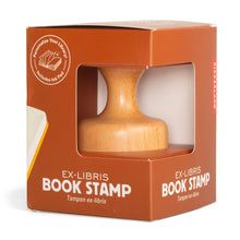 Load image into Gallery viewer, This image shows the retail packaging for an "Ex-Libris Book Stamp" by Kikkerland, housed in a square, warm-brown cardboard box with a large cutout window that displays a smooth, light-colored wooden stamp handle inside. The front of the box features bold white text labeling the product and an upper-left circular graphic that reads "Personalize Your Library! Includes Ink Pad" next to an illustration of an open book.
