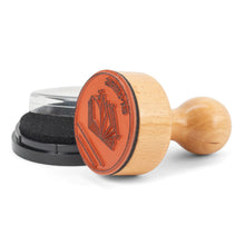 Load image into Gallery viewer, This image shows the unpackaged book stamp lying on its side against a white background, showcasing its round, orange rubber stamping surface carved with an open-book design and mirrored "EX-LIBRIS" text. Resting just behind the smooth wooden handle is a small, round black ink pad with its clear plastic lid removed.
