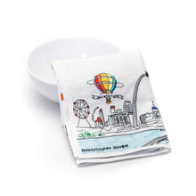 Load image into Gallery viewer, A folded white dish towel, resting against a plain white ceramic bowl, features colorful embroidery depicting famous St. Louis landmarks such as the Gateway Arch, Busch Stadium, a Ferris wheel, a hot air balloon, and the Mississippi River.
