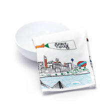 Load image into Gallery viewer, A folded white dish towel, propped against a plain white bowl, features colorful embroidery of the St. Louis skyline, a airplane banner reading "Spirit of St. Louis," the Mississippi River, and surrounding city landmarks.
