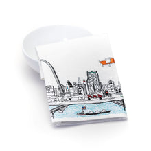 Load image into Gallery viewer, A folded white dish towel, resting against a plain white bowl, features detailed colorful embroidery showcasing St. Louis landmarks including the Gateway Arch, city skyline buildings, a horse-drawn carriage, an orange airplane, and a riverboat on the Mississippi River.

