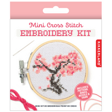 Load image into Gallery viewer, This retail package features a white card with a soft pink top border that showcases a finished "Mini Cross Stitch Embroidery Kit" project. The image on the packaging shows a small circular wooden hoop framing a stitched cross-stitch design of a dark grey tree branch blooming with vibrant pink cherry blossoms.
