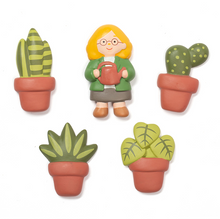 Load image into Gallery viewer, five individual, sculpted refrigerator magnets are displayed arranged in a loose cluster. In the center is a smiling woman with glasses and bright yellow hair holding a small watering can, completely surrounded by four unique potted houseplant magnets including a cactus, a snake plant, and two leafy green varieties in terracotta pots.
