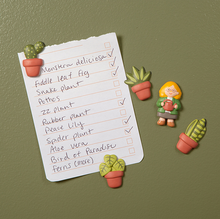 Load image into Gallery viewer, Set against a textured, olive-green surface, a handwritten checklist of plant names is held up by several of the colorful, 3D houseplant magnets. The tiny, smiling "Plant Lady" magnet and another potted plant magnet sit directly on the green background to the right of the note, completing the charming, organized scene.
