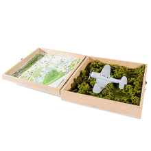 Load image into Gallery viewer, An open hinged wooden display case containing a miniature grey model airplane set on green moss on one side, and an illustrated map in the lid.

