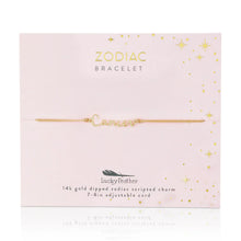 Load image into Gallery viewer, This image shows a gold "Cancer" script bracelet mounted flat against a soft, pale pink square card embellished with tiny metallic gold stars and dots. The minimalist bracelet features a delicate cursive nameplate charm dipped in 14k gold, threaded onto a thin, adjustable tan cord.
