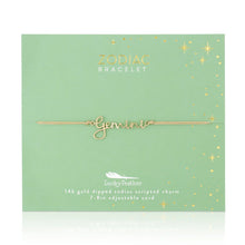 Load image into Gallery viewer, This image shows a gold "Gemini" script bracelet mounted flat against a sage green square card decorated with tiny metallic gold stars and dots. The minimalist bracelet features a delicate cursive nameplate charm dipped in 14k gold, threaded onto a thin, adjustable tan cord.
