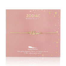 Load image into Gallery viewer, This image shows a gold "Libra" script bracelet mounted flat against a dusty rose square card adorned with tiny metallic gold stars and dots. The minimalist bracelet features a delicate cursive nameplate charm dipped in 14k gold, threaded onto a thin, adjustable tan cord.

