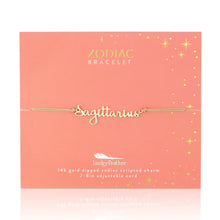 Load image into Gallery viewer, This image shows a gold "Sagittarius" script bracelet mounted flat against a vibrant coral-orange square card decorated with tiny metallic gold stars and dots. The minimalist bracelet features a delicate cursive nameplate charm dipped in 14k gold, threaded onto a thin, adjustable tan cord.
