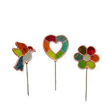 Load image into Gallery viewer, a set of three decorative, stained-glass style plant stakes nestled among green foliage and yellow flowers. Standing on slender silver rods, the stakes feature translucent, multi-colored glass panels framed in a copper-toned border, shaped respectively as a bird in flight, an open heart, and a five-petaled flower.
