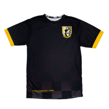 Load image into Gallery viewer, This short-sleeved black soccer jersey, trimmed with yellow cuffs at the sleeves. On the left chest sits a shield-shaped crest labeled "WCHOF FC" with a king chess piece silhouette, while the lower hem displays subtle checkered dark gray patterning along with "WORLD CHESS HALL OF FAME" printed in yellow.
