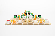 Load image into Gallery viewer, A eye-level, front-facing view shows the entire sushi chess set arranged on a bamboo mat board, featuring hand roll kings and queens standing on small stands.
