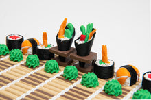 Load image into Gallery viewer, A close-up side angle focuses on the detailed dark-sided sushi pieces lining the bamboo mat, with wasabi ginger pawns lined up in front.
