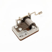 Load image into Gallery viewer, This image shows a small, exposed mechanical music box mechanism sitting at an angle against a solid white background. It features a silver metal base holding a textured rotating drum with tiny pins, a hand-crank arm with a cylindrical handle on the right, and a metal comb with fine teeth on the left that creates the musical notes.
