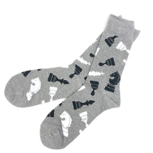 Load image into Gallery viewer,  A pair of grey crew-length socks displayed side-by-side against a plain white background. The fabric is patterned with stylized, graphic silhouettes of black and white chess pieces, including knights, rooks, kings, and pawns, scattered across the entire surface.
